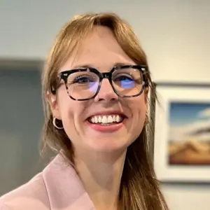 A woman with long, light brown hair and tortoiseshell glasses smiles broadly. She wears gold hoop earrings and a pale pink blazer. Blurred indoor background with a framed painting.